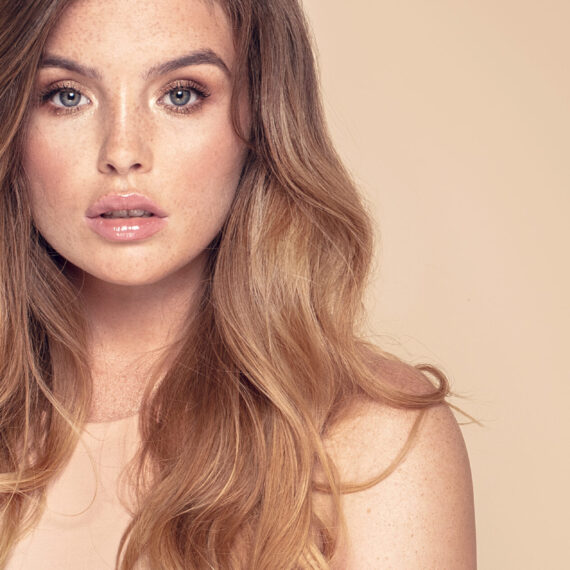 Portrait of beautiful young natural woman with freckles on her face. Girl with long wavy hair looking at camera. Beige studio background.